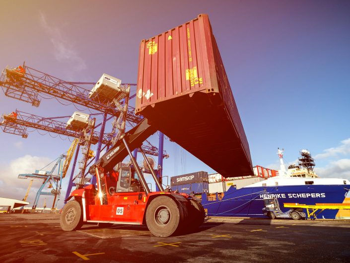A shipping photograph of a reachstacker forklift handling a container at Hull ABP container terminal with a ship and cranes in the background.